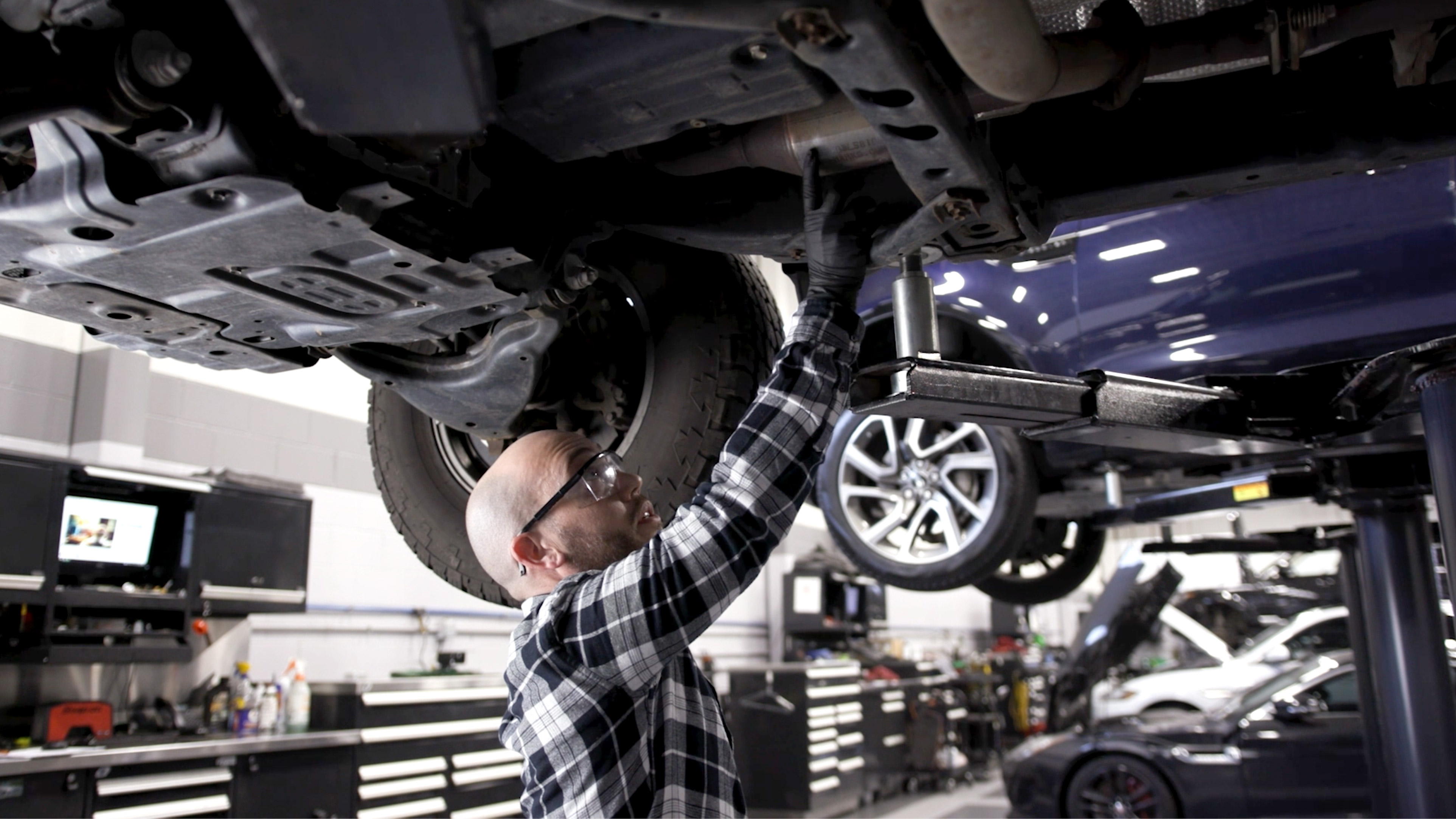 Man in a plaid shirt and safety glasses touching a catalytic converter under a lifter car.